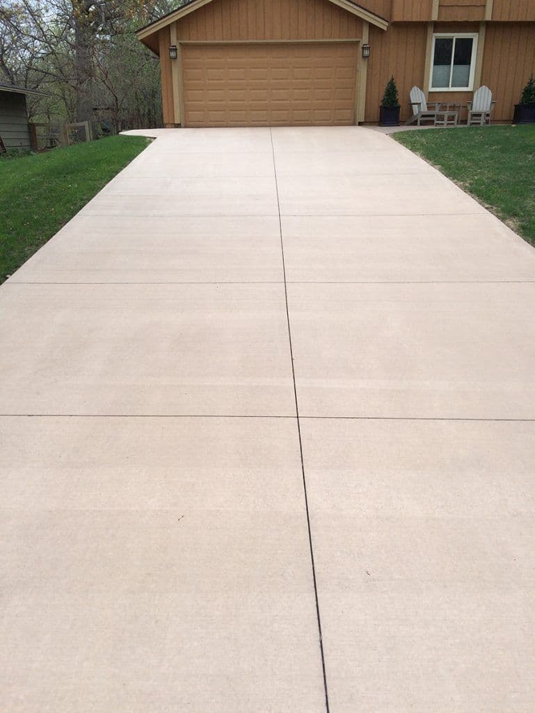 Concrete driveway leading to a house with a brown garage door and lawn chairs nearby.